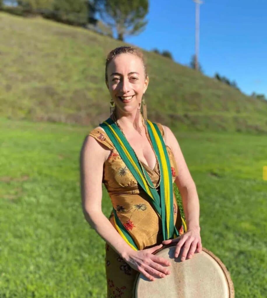 Carrie standing in front of a green hill with a drum hanging around her neck